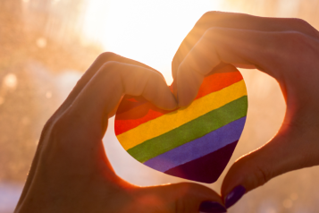 Two hands together in the shape of a heart holding a multi coloured heart in the sunshine.